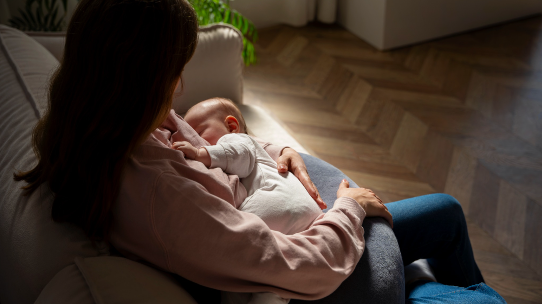A mother cradles her sleeping newborn while sitting in a softly lit living room.